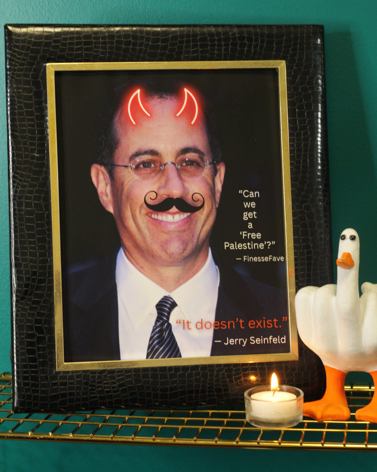 A framed novelty portrait print with a black crocodile-embossed frame and gold inner border, showing a man in a suit with devil horns and a mustache, plus quotes by Jerry Seinfeld. The frame is displayed on a wire shelf against a teal wall, next to a lit candle and a white goose figurine.